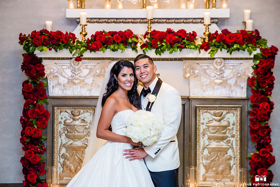 San diego glamorous wedding at the us grant hotel bride strapless ball gown with a tulle skirt and sweetheart neckline and a crystal belt with a long veil and groom white tuxedo jacket with a black shawl and black pants with a black bow tie and white floral boutonniere hugging and bride holding white floral bridal bouquet