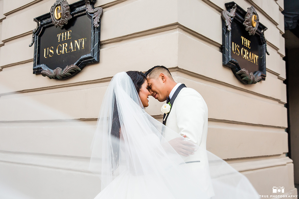 San diego glamorous wedding at the us grant hotel bride strapless ball gown with a tulle skirt and sweetheart neckline and a crystal belt with a long veil and groom white tuxedo jacket with a black shawl and black pants with a black bow tie and white floral boutonniere hugging outside hotel