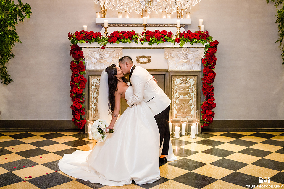 San diego glamorous wedding at the us grant hotel bride strapless ball gown with a tulle skirt and sweetheart neckline and a crystal belt with a long veil and groom white tuxedo jacket with a black shawl and black pants with a black bow tie and white floral boutonniere hugging and kissing bride holding white floral bridal bouquet