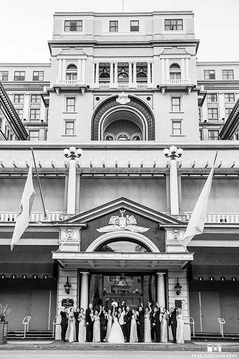 San diego glamorous wedding at the us grant hotel bride strapless ball gown with a tulle skirt and sweetheart neckline and a crystal belt with a long veil and groom white tuxedo jacket with a black shawl and black pants with a black bow tie and white floral boutonniere with wedding party bridesmaids long gold dresses and groomsmen black tuxedos cheering black and white photo