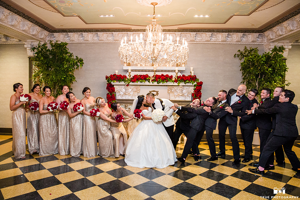 San diego glamorous wedding at the us grant hotel bride strapless ball gown with a tulle skirt and sweetheart neckline and a crystal belt with a long veil and groom white tuxedo jacket with a black shawl and black pants with a black bow tie and white floral boutonniere kissing with wedding party bridesmaids long gold dresses with red floral bouquets and groomsmen black tuxedos with black bow ties