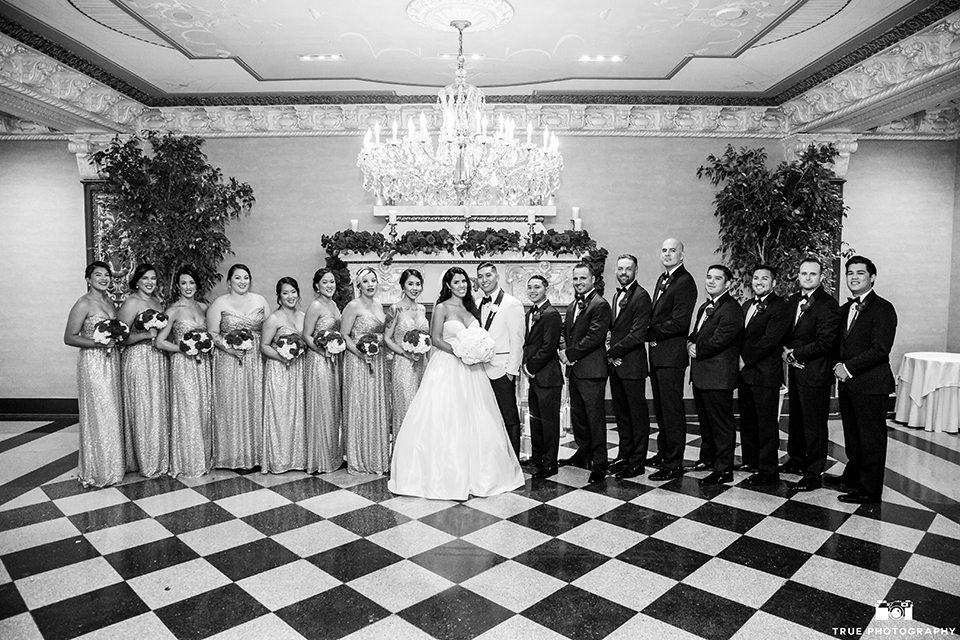 San diego glamorous wedding at the us grant hotel bride strapless ball gown with a tulle skirt and sweetheart neckline and a crystal belt with a long veil and groom white tuxedo jacket with a black shawl and black pants with a black bow tie and white floral boutonniere with wedding party bridesmaids long gold dresses with red floral bouquets and groomsmen black tuxedos with black bow ties