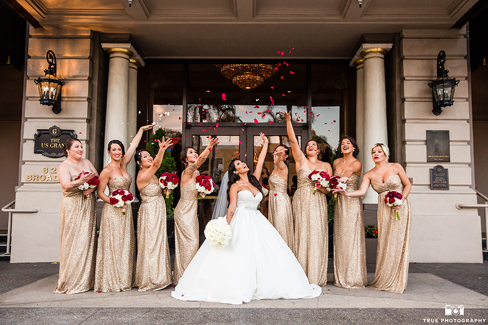 San diego glamorous wedding at the us grant hotel bride strapless ball gown with a tulle skirt and sweetheart neckline and a crystal belt with a long veil holding white floral bridal bouquet with bridesmaids long gold dresses and red floral bouquets cheering