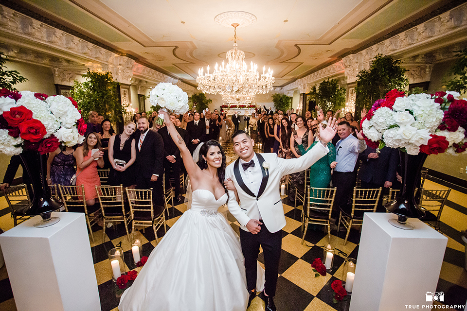 San diego glamorous wedding at the us grant hotel bride strapless ball gown with a tulle skirt and sweetheart neckline and a crystal belt with a long veil and groom white tuxedo jacket with a black shawl and black pants with a black bow tie and white floral boutonniere cheering after ceremony