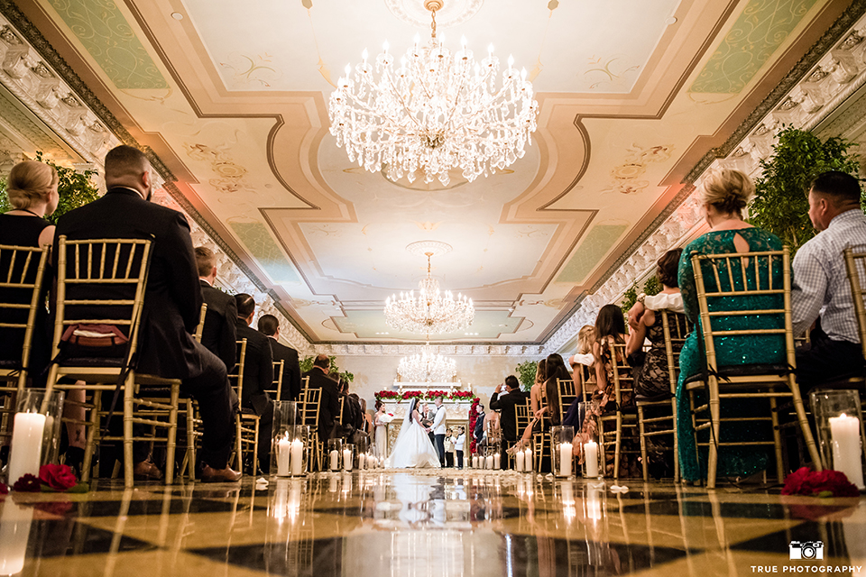 San diego glamorous wedding at the us grant hotel bride strapless ball gown with a tulle skirt and sweetheart neckline and a crystal belt with a long veil and groom white tuxedo jacket with a black shawl and black pants with a black bow tie and white floral boutonniere holding hands during ceremony