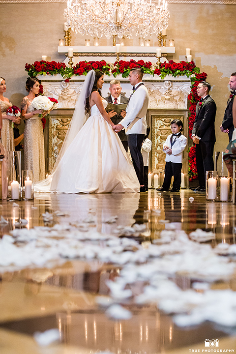 San diego glamorous wedding at the us grant hotel bride strapless ball gown with a tulle skirt and sweetheart neckline and a crystal belt with a long veil and groom white tuxedo jacket with a black shawl and black pants with a black bow tie and white floral boutonniere holding hands during ceremony