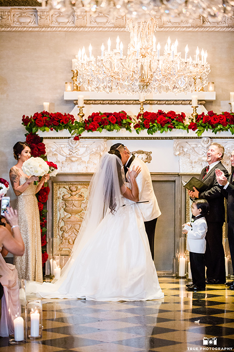 San diego glamorous wedding at the us grant hotel bride strapless ball gown with a tulle skirt and sweetheart neckline and a crystal belt with a long veil and groom white tuxedo jacket with a black shawl and black pants with a black bow tie and white floral boutonniere kissing and hugging during ceremony