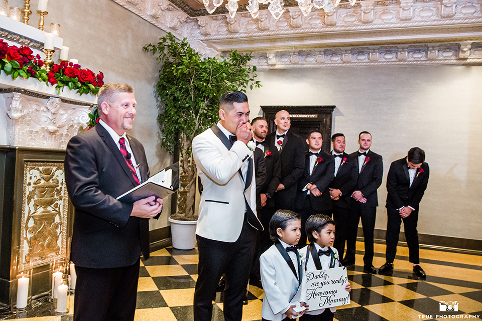 San diego glamorous wedding at the us grant hotel groom white tuxedo with black shawl lapel and black tuxedo pants with a white dress shirt and black bow tie with a white floral boutonniere crying watching bride walk down the aisle during ceremony