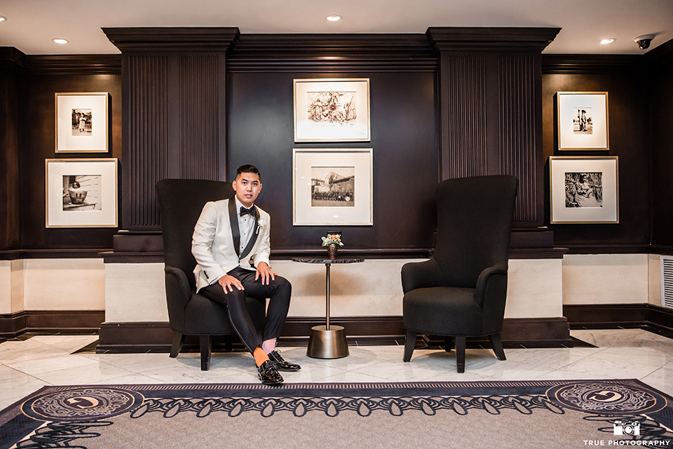 San diego glamorous wedding at the us grant hotel groom white tuxedo with black shawl lapel and black tuxedo pants with a white dress shirt and black bow tie with a white floral boutonniere sitting in black chair