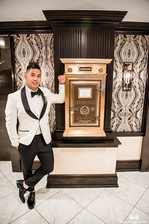 San diego glamorous wedding at the us grant hotel groom white tuxedo with black shawl lapel and black tuxedo pants with a white dress shirt and black bow tie with a white floral boutonniere standing with hands in pockets