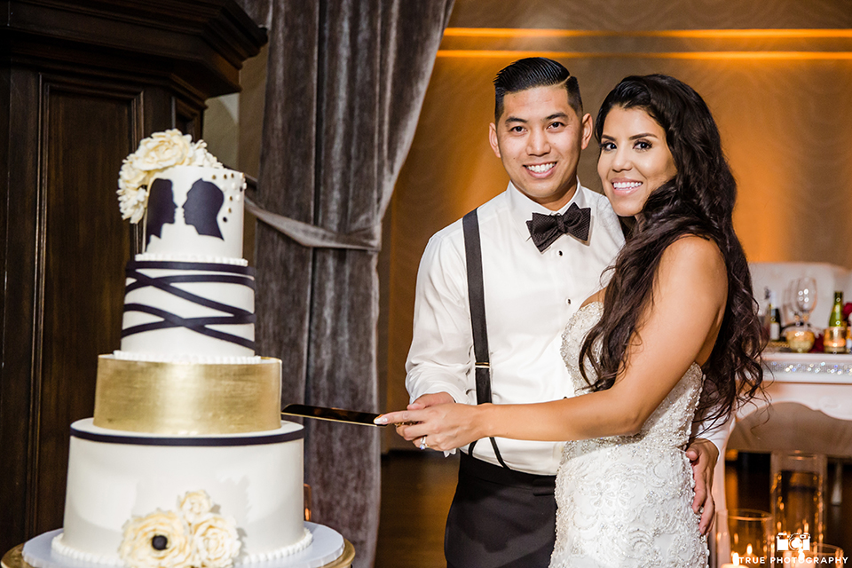 San diego glamorous wedding at the us grant hotel bride strapless ball gown with a tulle skirt and sweetheart neckline and a crystal belt with a long veil and groom white tuxedo jacket with a black shawl and black pants with a black bow tie and white floral boutonniere cutting cake