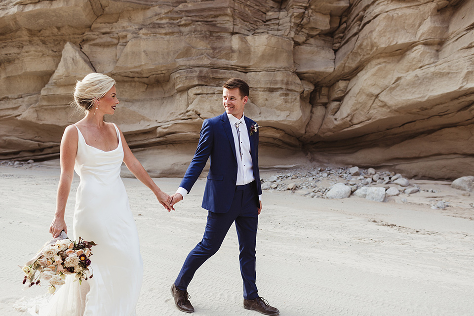 Anza-Borrego-styled-shoot-bride-and-groom-holding-hands-walking-bride-in-white-silk-bridal-gown-with-open-back-and-thin-straps-groom-in-colbalt-blue-suit-with-chocolate-brown-shoes-and-bolo-tie