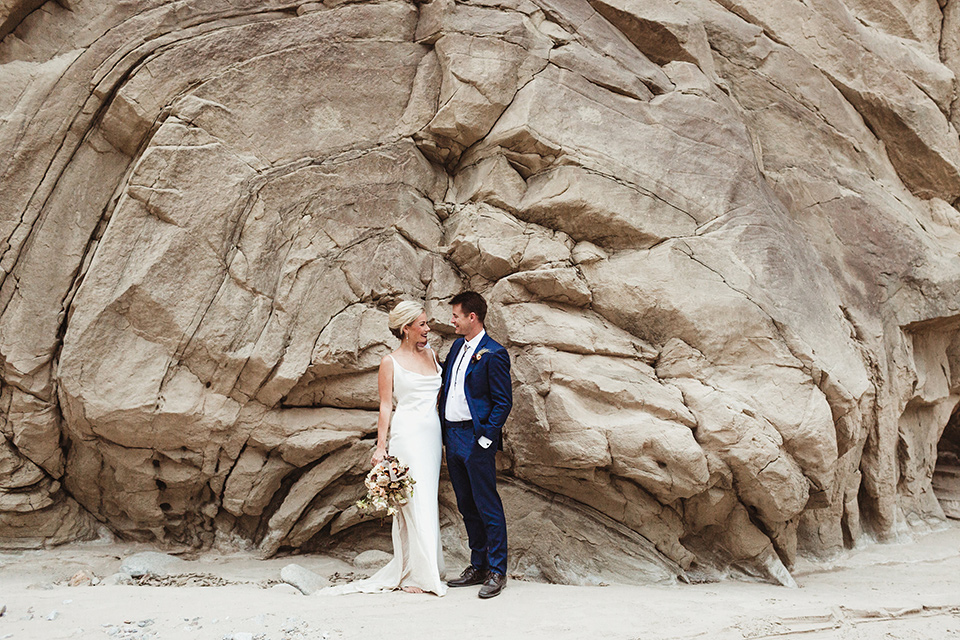 Anza-Borrego-styled-shoot-bride-and-groom-in-front-of-rocks-bride-in-white-silk-bridal-gown-with-open-back-and-thin-straps-groom-in-colbalt-blue-suit-with-chocolate-brown-shoes-and-bolo-tie