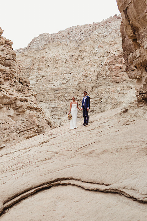 Anza-Borrego-styled-shoot-bride-and-groom-on-slanted-rocks-bride-in-white-silk-bridal-gown-with-open-back-and-thin-straps-groom-in-colbalt-blue-suit-with-chocolate-brown-shoes-and-bolo-tie