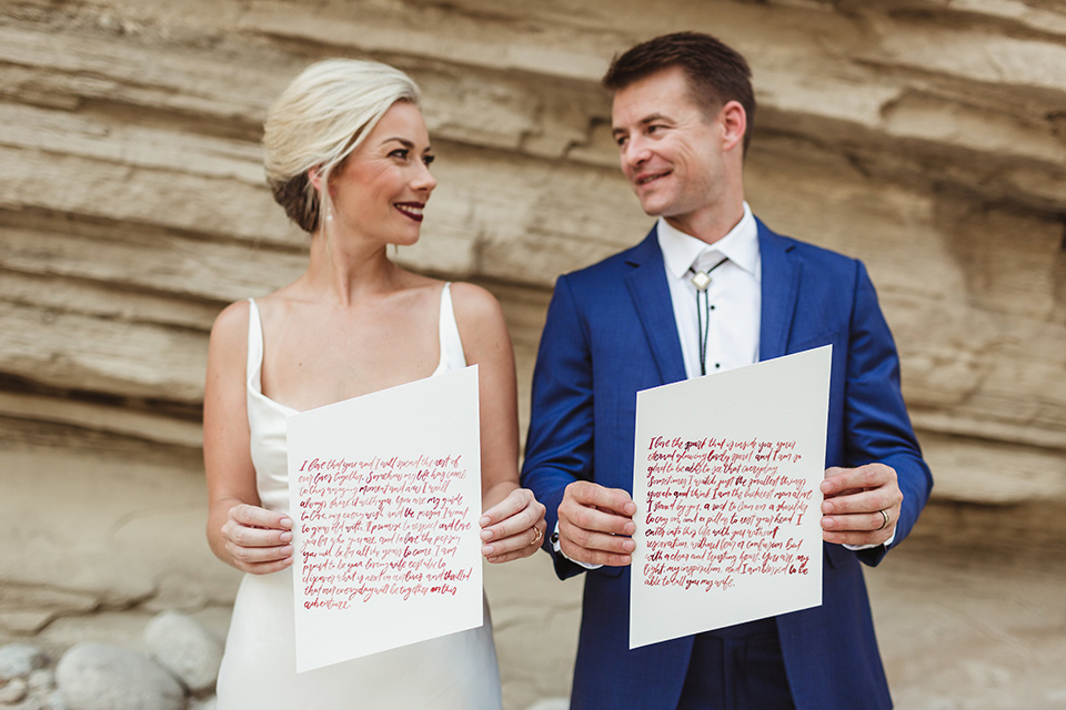 Anza-Borrego-styled-shoot-holding-calligraphy-papers-bride-in-white-silk-bridal-gown-with-open-back-and-thin-straps-groom-in-colbalt-blue-suit-with-chocolate-brown-shoes-and-bolo-tie