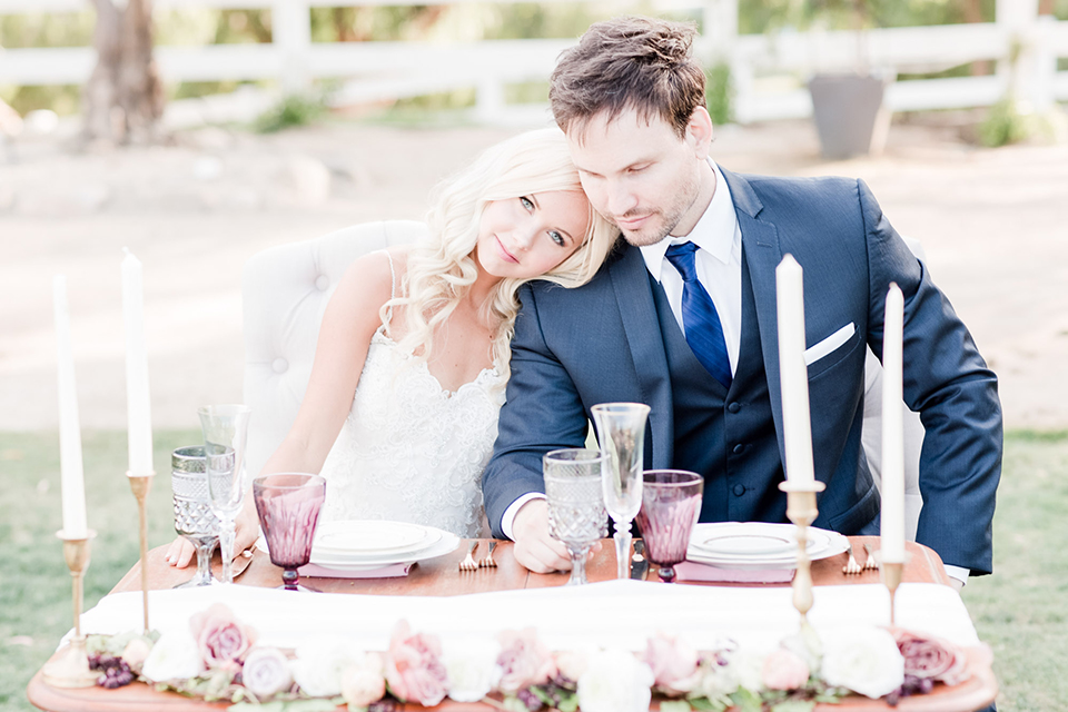 Los angeles outdoor wedding at brookview ranch bride form fitting gown with lace detail on bodice and straps with a plunging neckline and groom navy blue notch lapel suit with a matching vest and white dress shirt with a long blue tie sitting at table