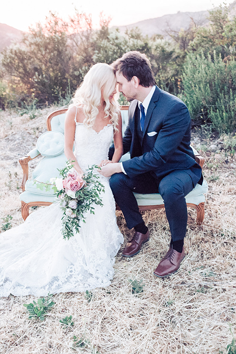 Los angeles outdoor wedding at brookview ranch bride form fitting gown with lace detail on bodice and straps with a plunging neckline and groom navy blue notch lapel suit with a matching vest and white dress shirt with a long blue tie sitting on couch hugging bride holding white and pink floral bridal bouquet