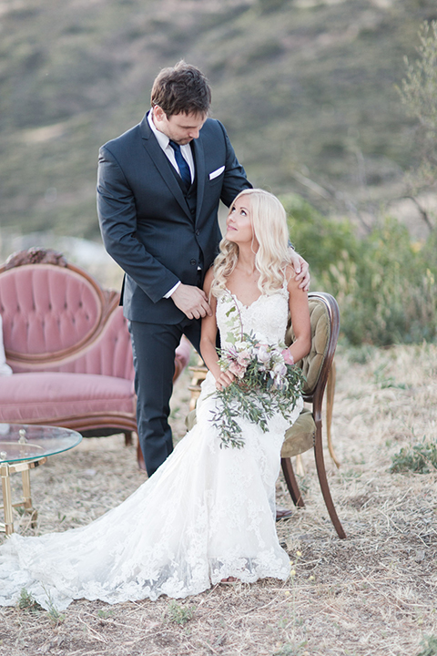 Los angeles outdoor wedding at brookview ranch bride form fitting gown with lace detail on bodice and straps with a plunging neckline and groom navy blue notch lapel suit with a matching vest and white dress shirt with a long blue tie sitting on chair and bride holding white and pink floral bridal bouquet