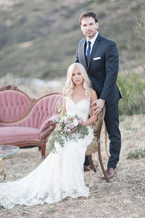 Los angeles outdoor wedding at brookview ranch bride form fitting gown with lace detail on bodice and straps with a plunging neckline and groom navy blue notch lapel suit with a matching vest and white dress shirt with a long blue tie sitting in chair and bride holding white and pink floral bridal bouquet