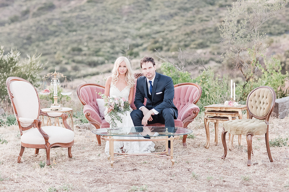Los angeles outdoor wedding at brookview ranch bride form fitting gown with lace detail on bodice and straps with a plunging neckline and groom navy blue notch lapel suit with a matching vest and white dress shirt with a long blue tie sitting on pink couch bride holding white and pink floral bridal bouquet