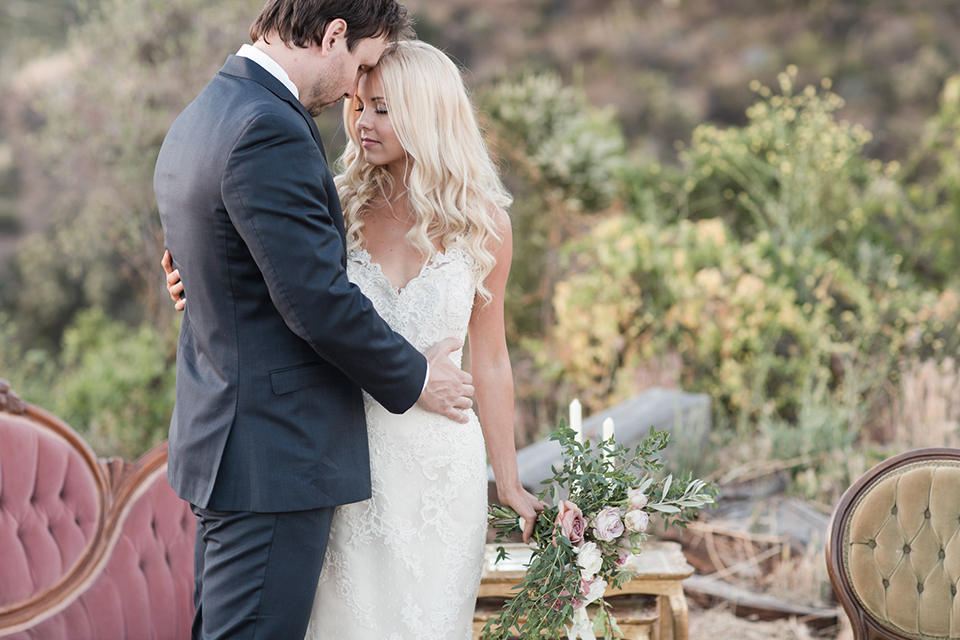 Los angeles outdoor wedding at brookview ranch bride form fitting gown with lace detail on bodice and straps with a plunging neckline and groom navy blue notch lapel suit with a matching vest and white dress shirt with a long blue tie hugging and bride holding white and pink floral bridal bouquet