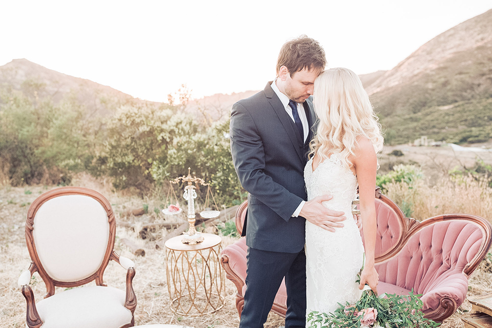 Los angeles outdoor wedding at brookview ranch bride form fitting gown with lace detail on bodice and straps with a plunging neckline and groom navy blue notch lapel suit with a matching vest and white dress shirt with a long blue tie standing and hugging bride holding white and pink floral bridal bouquet