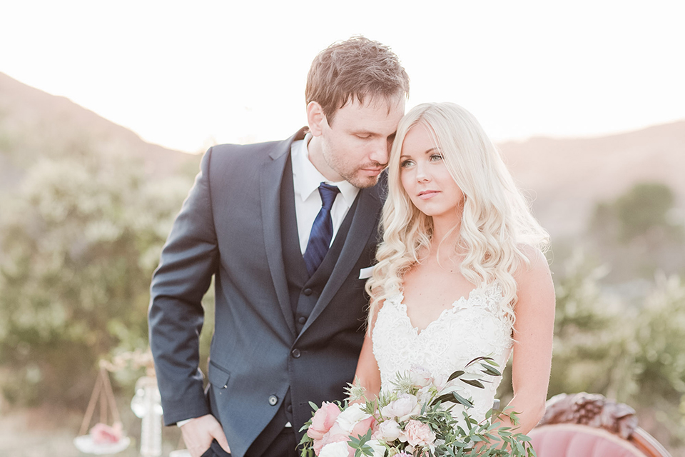 Los angeles outdoor wedding at brookview ranch bride form fitting gown with lace detail on bodice and straps with a plunging neckline and groom navy blue notch lapel suit with a matching vest and white dress shirt with a long blue tie hugging and bride holding white and pink floral bridal bouquet