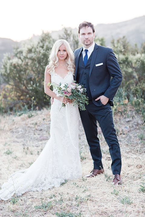 Los angeles outdoor wedding at brookview ranch bride form fitting gown with lace detail on bodice and straps with a plunging neckline and groom navy blue notch lapel suit with a matching vest and white dress shirt with a long blue tie standing and bride holding white and pink floral bridal bouquet