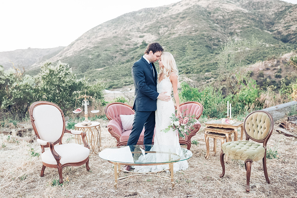 Los angeles outdoor wedding at brookview ranch bride form fitting gown with lace detail on bodice and straps with a plunging neckline and groom navy blue notch lapel suit with a matching vest and white dress shirt with a long blue tie hugging and bride holding white and pink floral bridal bouquet