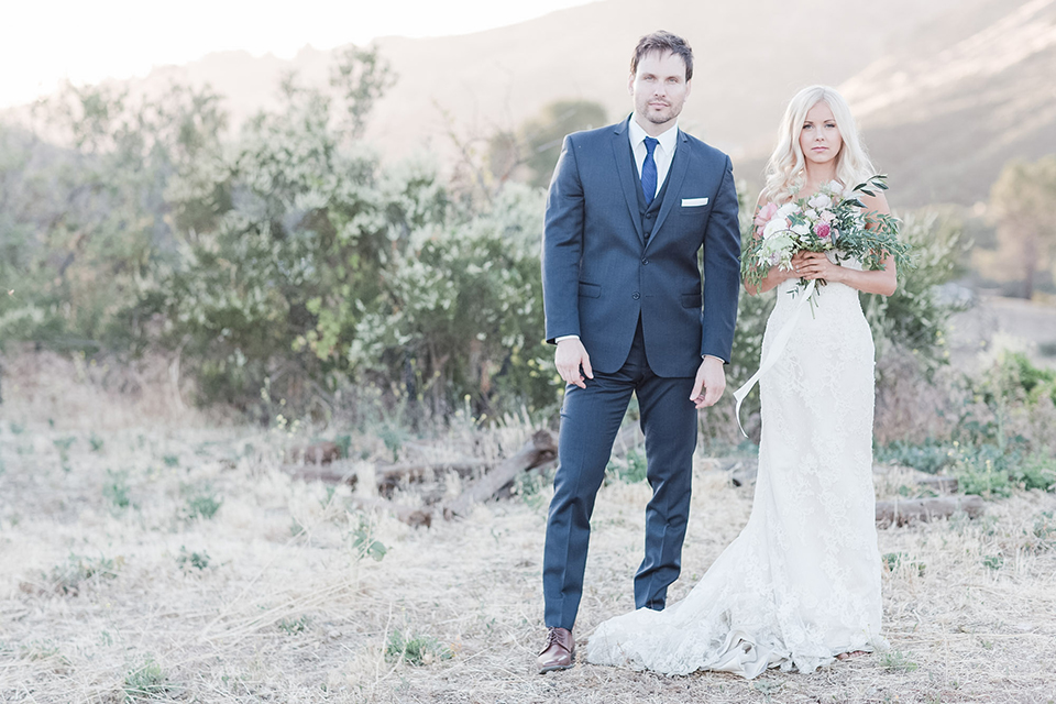 Los angeles outdoor wedding at brookview ranch bride form fitting gown with lace detail on bodice and straps with a plunging neckline and groom navy blue notch lapel suit with a matching vest and white dress shirt with a long blue tie holding hands and bride holding white and pink floral bridal bouquet