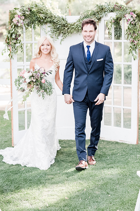 Los angeles outdoor wedding at brookview ranch bride form fitting gown with lace detail on bodice and straps with a plunging neckline and groom navy blue notch lapel suit with a matching vest and white dress shirt with a long blue tie holding hands and bride holding white and pink floral bridal bouquet