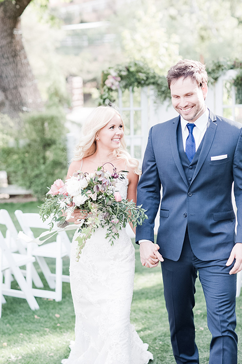 Los angeles outdoor wedding at brookview ranch bride form fitting gown with lace detail on bodice and straps with a plunging neckline and groom navy blue notch lapel suit with a matching vest and white dress shirt with a long blue tie holding hands and walking bride holding white and pink floral bridal bouquet