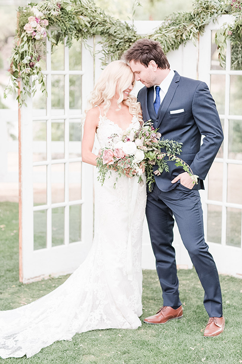 Los angeles outdoor wedding at brookview ranch bride form fitting gown with lace detail on bodice and straps with a plunging neckline and groom navy blue notch lapel suit with a matching vest and white dress shirt with a long blue tie hugging and bride holding white and pink floral bridal bouquet
