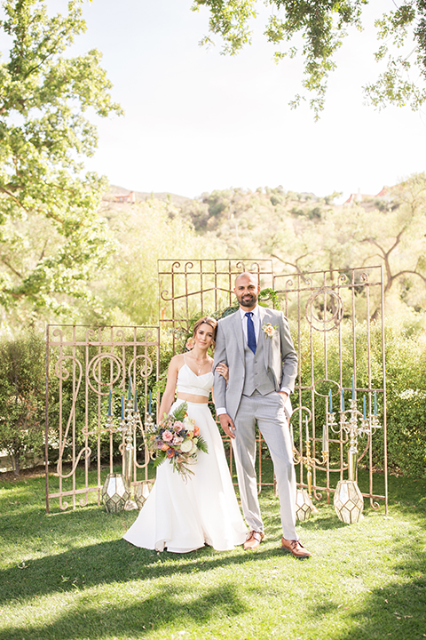 Los angeles outdoor wedding at brookview ranch bride form fitting gown with lace detail on bodice and straps with a plunging neckline and groom navy blue notch lapel suit with a matching vest and white dress shirt with a long blue tie hugging and bride holding white and pink floral bridal bouquet