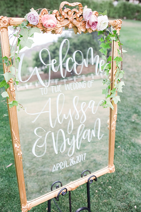 Los angeles outdoor wedding at brookview ranch ceremony set up on grass with white chairs and white window pane backdrop decor for altar and clear glass welcome sign with white calligraphy writing and greenery floral decor