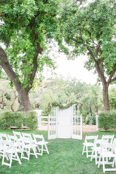 Los angeles outdoor wedding at brookview ranch ceremony set up on grass with white chairs and white window pane backdrop decor for altar and clear glass welcome sign with white calligraphy writing and greenery floral decor