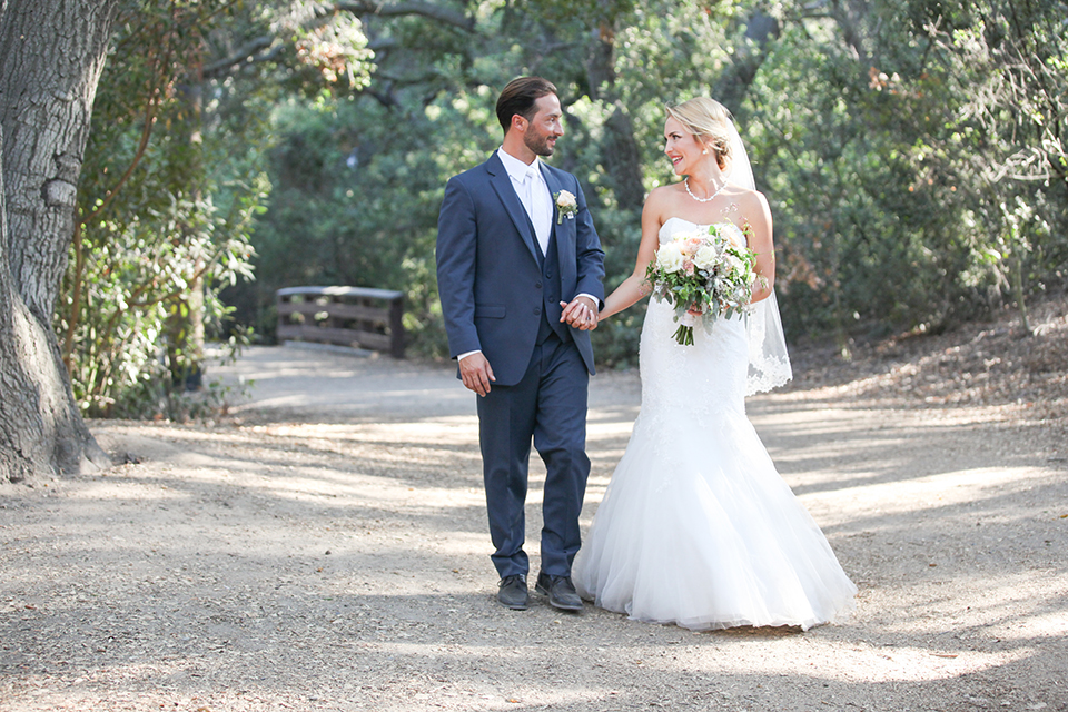 Orange county outdoor wedding at the oak canyon nature center bride form fitting mermaid style strapless gown with a crystal belt and sweetheart neckline with a long veil and groom slate blue notch lapel suit with a matching vest and white dress shirt with a long white tie and white floral boutonniere holding hands and bride holding white and green floral bridal bouquet