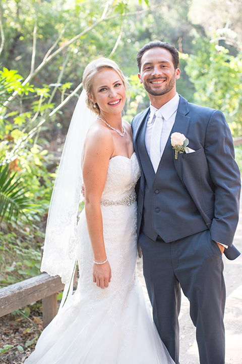 Orange county outdoor wedding at the oak canyon nature center bride form fitting mermaid style strapless gown with a crystal belt and sweetheart neckline with a long veil and groom slate blue notch lapel suit with a matching vest and white dress shirt with a long white tie and white floral boutonniere hugging