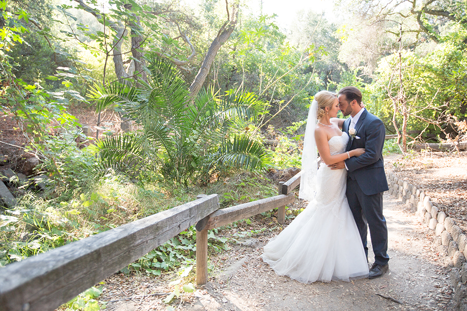 Orange county outdoor wedding at the oak canyon nature center bride form fitting mermaid style strapless gown with a crystal belt and sweetheart neckline with a long veil and groom slate blue notch lapel suit with a matching vest and white dress shirt with a long white tie and white floral boutonniere hugging