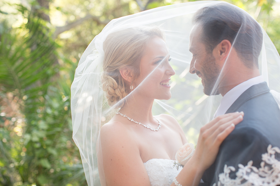 Orange county outdoor wedding at the oak canyon nature center bride form fitting mermaid style strapless gown with a crystal belt and sweetheart neckline with a long veil and groom slate blue notch lapel suit with a matching vest and white dress shirt with a long white tie and white floral boutonniere hugging under veil