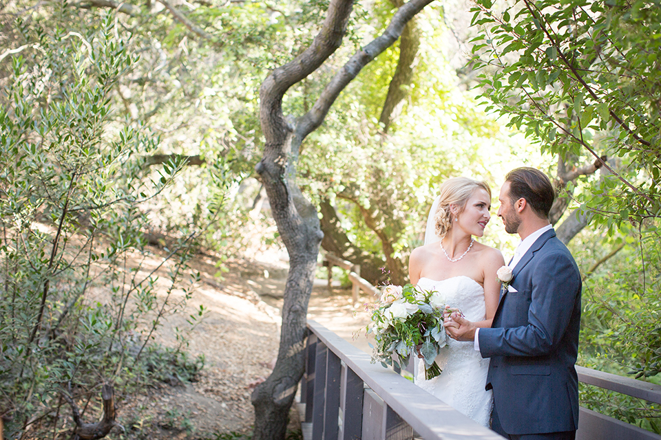 Orange county outdoor wedding at the oak canyon nature center bride form fitting mermaid style strapless gown with a crystal belt and sweetheart neckline with a long veil and groom slate blue notch lapel suit with a matching vest and white dress shirt with a long white tie and white floral boutonniere standing and hugging bride holding white and green floral bridal bouquet