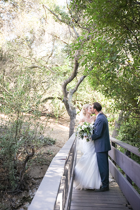 Orange county outdoor wedding at the oak canyon nature center bride form fitting mermaid style strapless gown with a crystal belt and sweetheart neckline with a long veil and groom slate blue notch lapel suit with a matching vest and white dress shirt with a long white tie and white floral boutonniere kissing bride holding white and green floral bridal bouquet