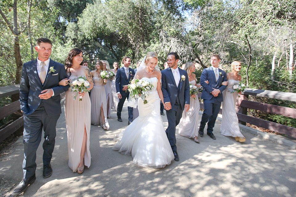 Orange county outdoor wedding at the oak canyon nature center bride form fitting mermaid style strapless gown with a crystal belt and sweetheart neckline with a long veil and groom slate blue notch lapel suit with a matching vest and white dress shirt with a long white tie and white floral boutonniere holding hands and walking with wedding party bridesmaids long grey dresses and groomsmen slate blue suits with long white ties