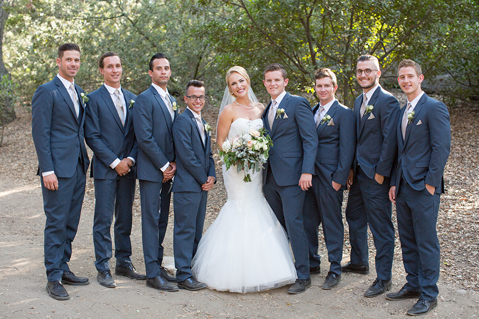 Orange county outdoor wedding at the oak canyon nature center bride form fitting mermaid style strapless gown with a crystal belt and sweetheart neckline with a long veil holding white and green floral bridal bouquet standing with groomsmen slate blue suits with long white ties