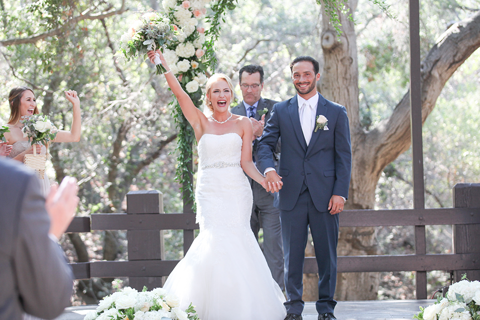 Orange county outdoor wedding at the oak canyon nature center bride form fitting mermaid style strapless gown with a crystal belt and sweetheart neckline with a long veil and groom slate blue notch lapel suit with a matching vest and white dress shirt with a long white tie and white floral boutonniere holding hands and cheering after ceremony