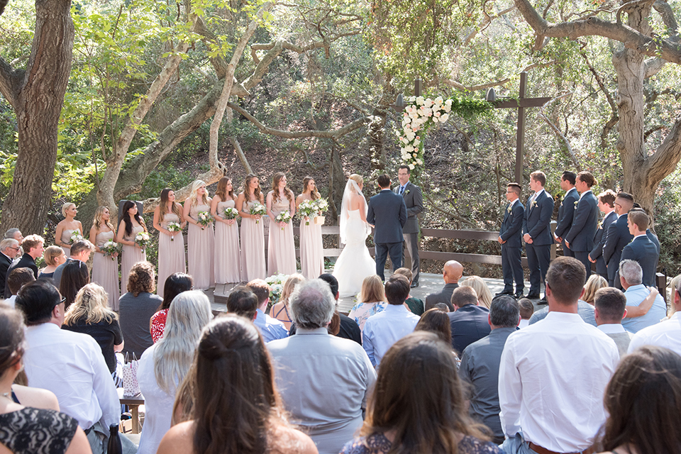 Orange county outdoor wedding at the oak canyon nature center bride form fitting mermaid style strapless gown with a crystal belt and sweetheart neckline with a long veil and groom slate blue notch lapel suit with a matching vest and white dress shirt with a long white tie and white floral boutonniere holding hands during ceremony