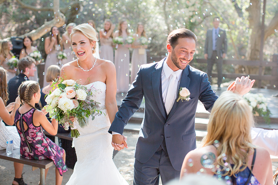 Orange county outdoor wedding at the oak canyon nature center bride form fitting mermaid style strapless gown with a crystal belt and sweetheart neckline with a long veil and groom slate blue notch lapel suit with a matching vest and white dress shirt with a long white tie and white floral boutonniere holding hands and walking down aisle after ceremony