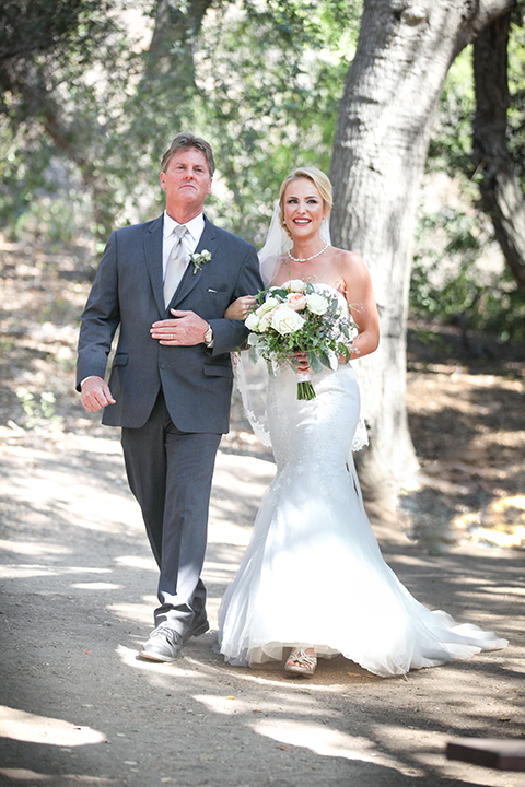 Orange county outdoor wedding at the oak canyon nature center bride form fitting mermaid style strapless gown with a crystal belt and sweetheart neckline with a long veil holding white and green floral bridal bouquet walking down the aisle with dad during ceremony