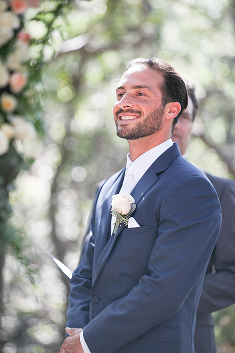 Orange county outdoor wedding at the oak canyon nature center groom slate blue notch lapel suit with a matching vest and white dress shirt with a long white tie and white floral boutonniere watching bride walk down the aisle during ceremony