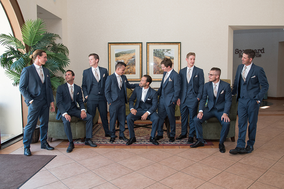 Orange county outdoor wedding at the oak canyon nature center groom slate blue notch lapel suit with a matching vest and white dress shirt with a long white tie and white floral boutonniere standing with groomsmen slate blue suits with long white ties sitting getting ready before ceremony